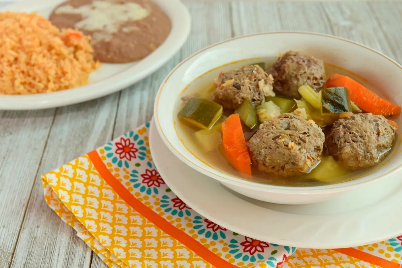Bowl of meatball soup with carrots and squash on a bright patterned napkin, rice and beans behind